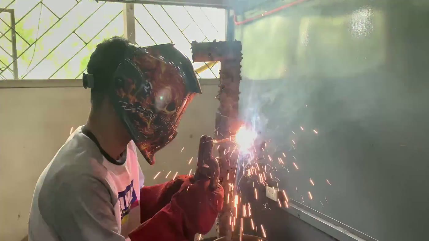 Welder in a flame-decorated helmet striking an arc on a steel bracket, sparks streaming across the Jacksonville shop floor